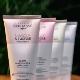 Cztery tubki maseczek do twarzy Byphasse w odcieniach bieli, różu i zieleni, ustawione rzędem na drewnianej powierzchni, z ciemnym, rozmyty tłem roślinnym.