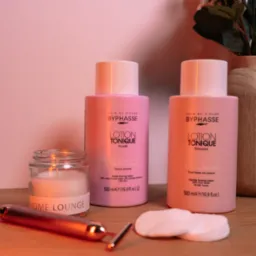 Dwa różowe toniki Byphasse obok świecy Home Lounge, wacików i masażera do twarzy, na drewnianym blacie, z wazonem kwiatów w tle. Ciepłe oświetlenie.