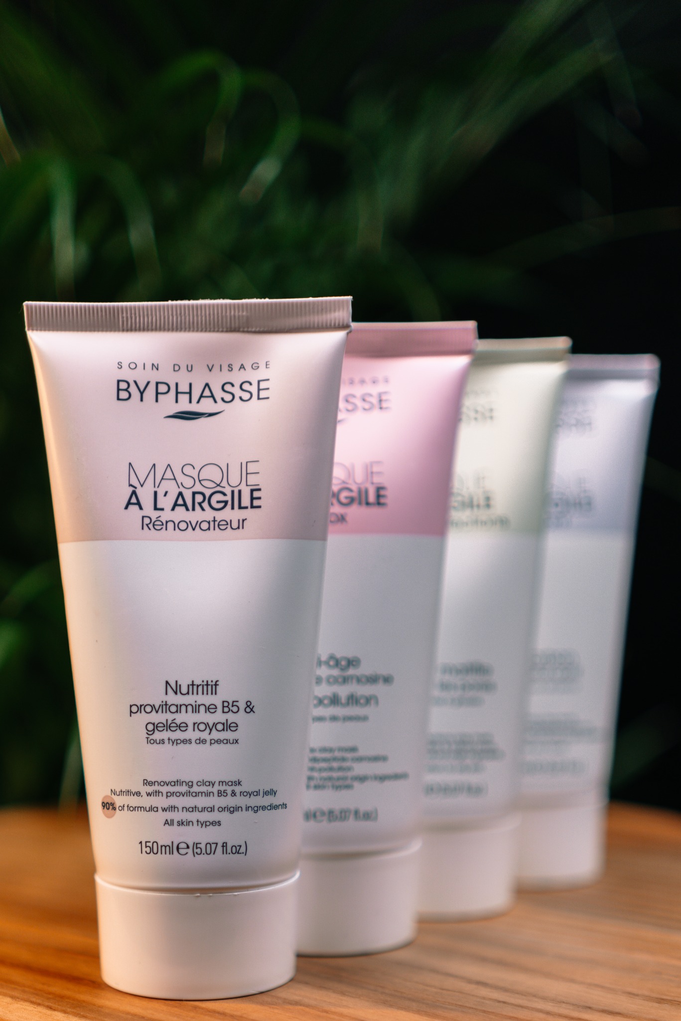 Cztery tubki maseczek do twarzy Byphasse w odcieniach bieli, różu i zieleni, ustawione rzędem na drewnianej powierzchni, z ciemnym, rozmyty tłem roślinnym.