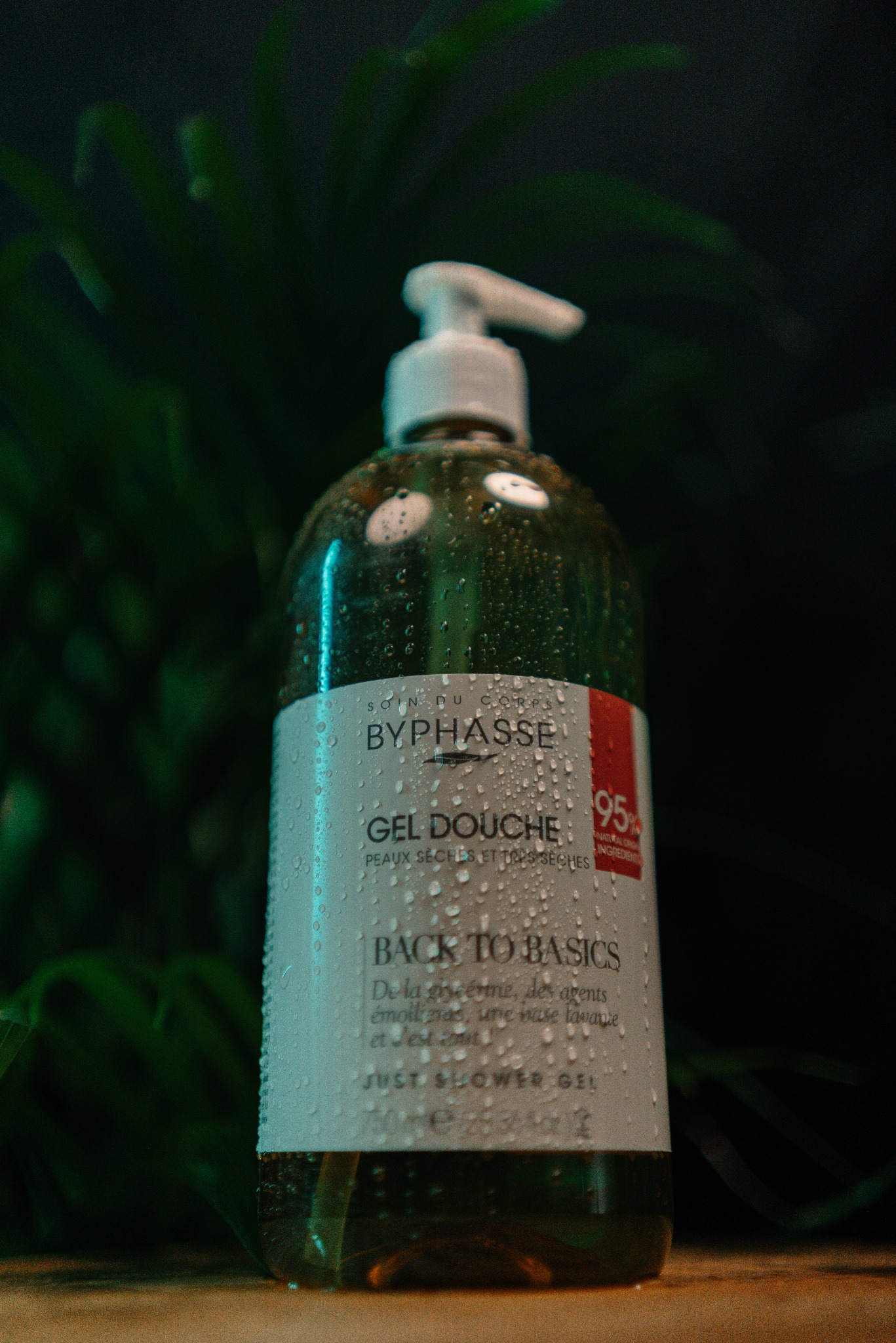 Butelka żelu pod prysznic Byphasse z kroplami wody na etykiecie, na rozmytym tle z zielonych liści. Produkt oświetlony zielonym światłem.