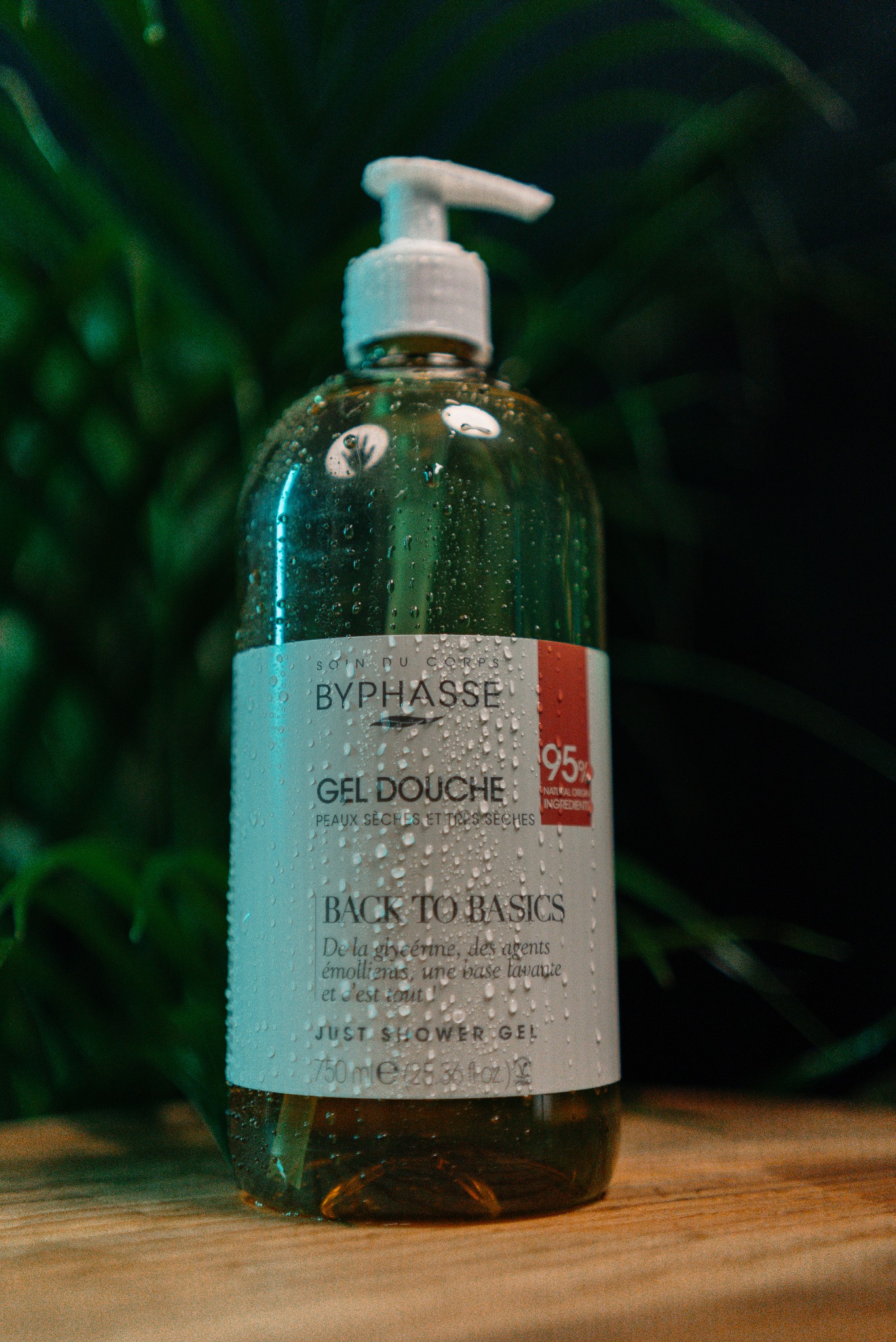 Butelka żelu pod prysznic Byphasse z pompką, pokryta kroplami wody. Widoczne zielone liście w tle. Produkt na drewnianej powierzchni w ciepłym oświetleniu.