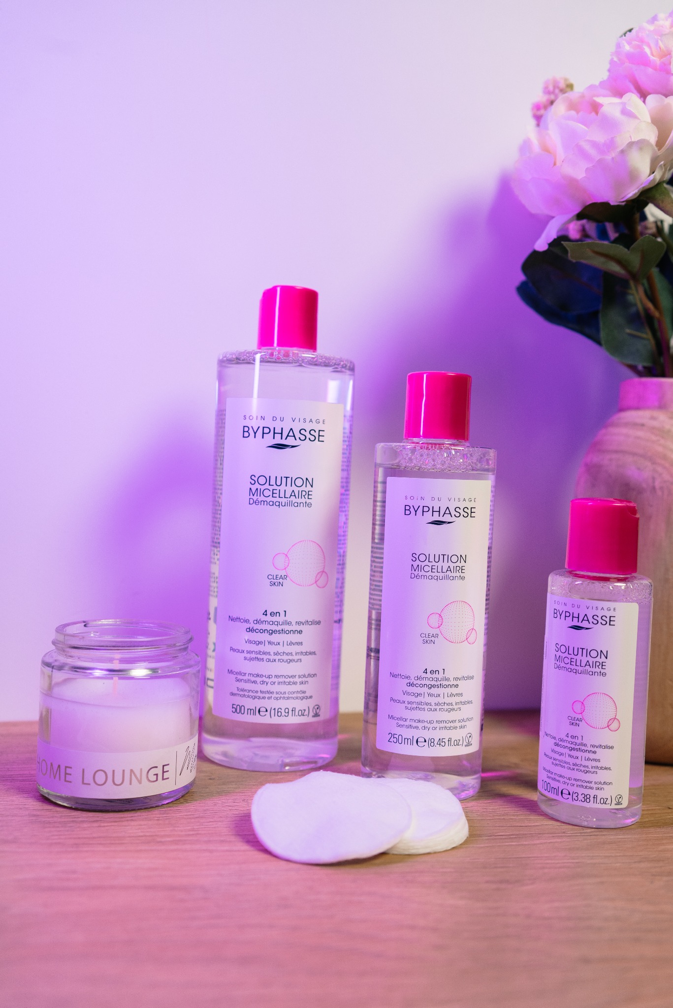 Trzy butelki płynu micelarnego Byphasse z różowymi nakrętkami, waciki kosmetyczne i świeca na drewnianym blacie, z kwiatami w tle.