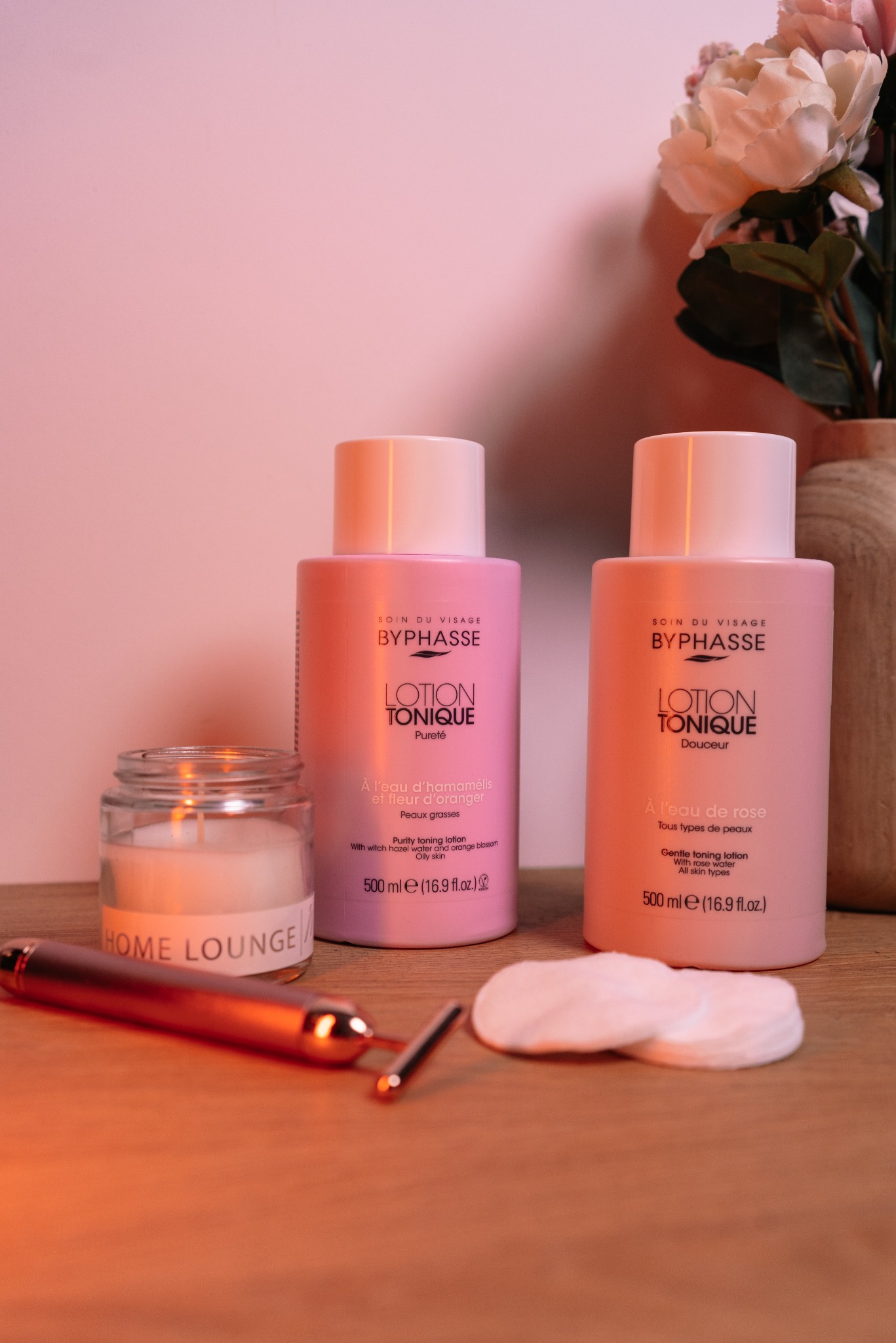 Dwa różowe toniki Byphasse obok świecy Home Lounge, wacików i masażera do twarzy, na drewnianym blacie, z wazonem kwiatów w tle. Ciepłe oświetlenie.