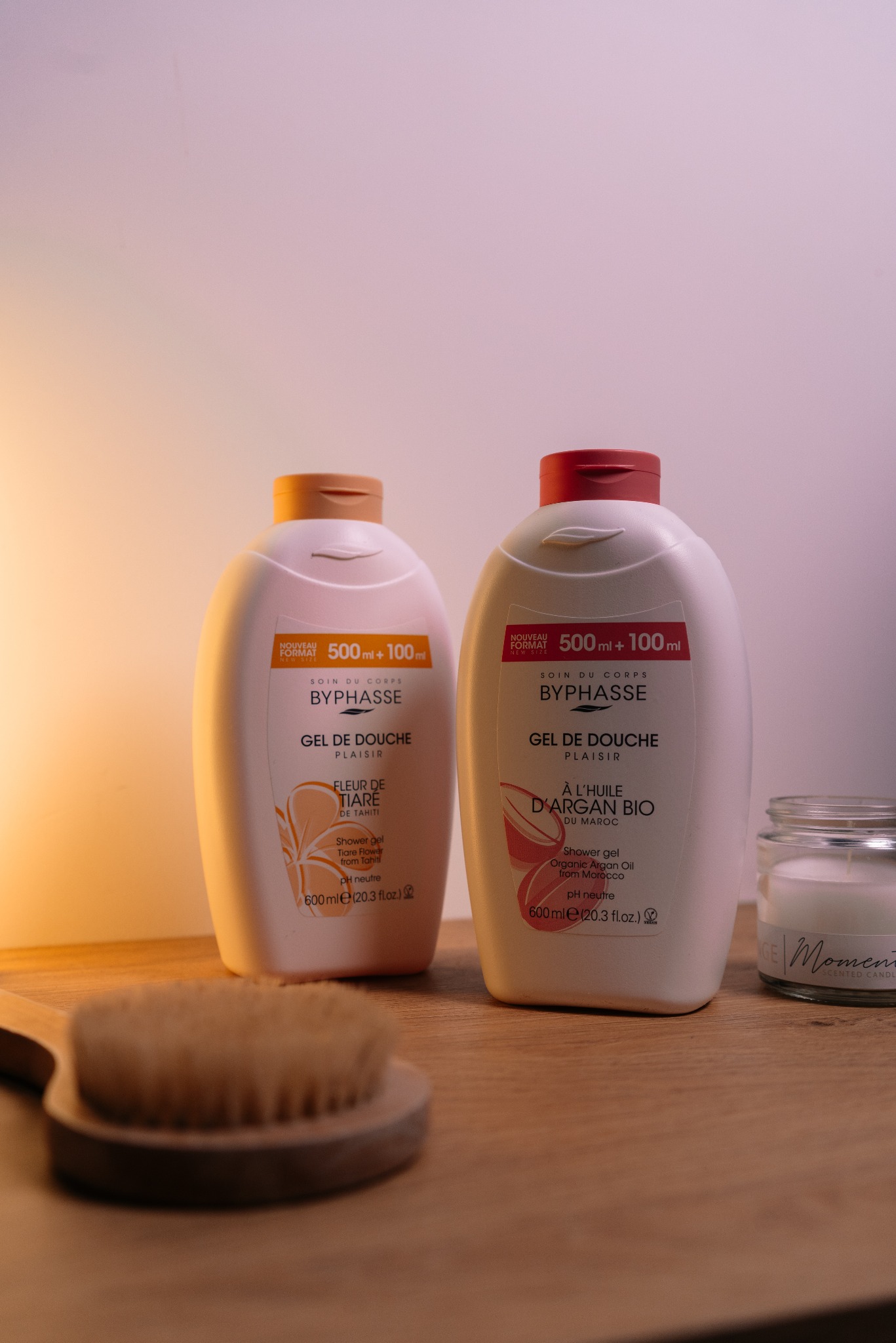 Dwa białe żele pod prysznic Byphasse (500ml+100ml) z brązowymi i różowymi zakrętkami, obok szczotka do masażu i świeca na drewnianym blacie, ciepłe oświetlenie.