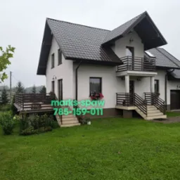 Dom z brązowymi balustradami balkonowymi i tarasowymi, widocznymi zieleń trawnika i kwiaty w oknach. Balustrady metalowe o prostej konstrukcji, dopasowane kolorystycznie do dachu.