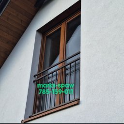 Łukasz Marks Usługi Spawalnicze - Balkon z brązową, metalową balustradą z pionowymi prętami, zamontowaną przy oknie w budynku z drewnianym podbitką. Widok z dołu, z lekkim kątem.