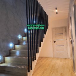 Łukasz Marks Usługi Spawalnicze - Nowoczesne schody z drewnianymi stopniami, podświetleniem LED i balustradą z pionowych, czarnych prętów. Ściana o fakturze betonu. Widok na korytarz z drewnianą podłogą i drzwiami.