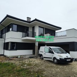 Łukasz Marks Usługi Spawalnicze - Nowoczesny dom z balkonami z balustradami szklanymi i czarnymi ramami okiennymi, biała bryła budynku z czarnymi akcentami, obok biały samochód dostawczy.