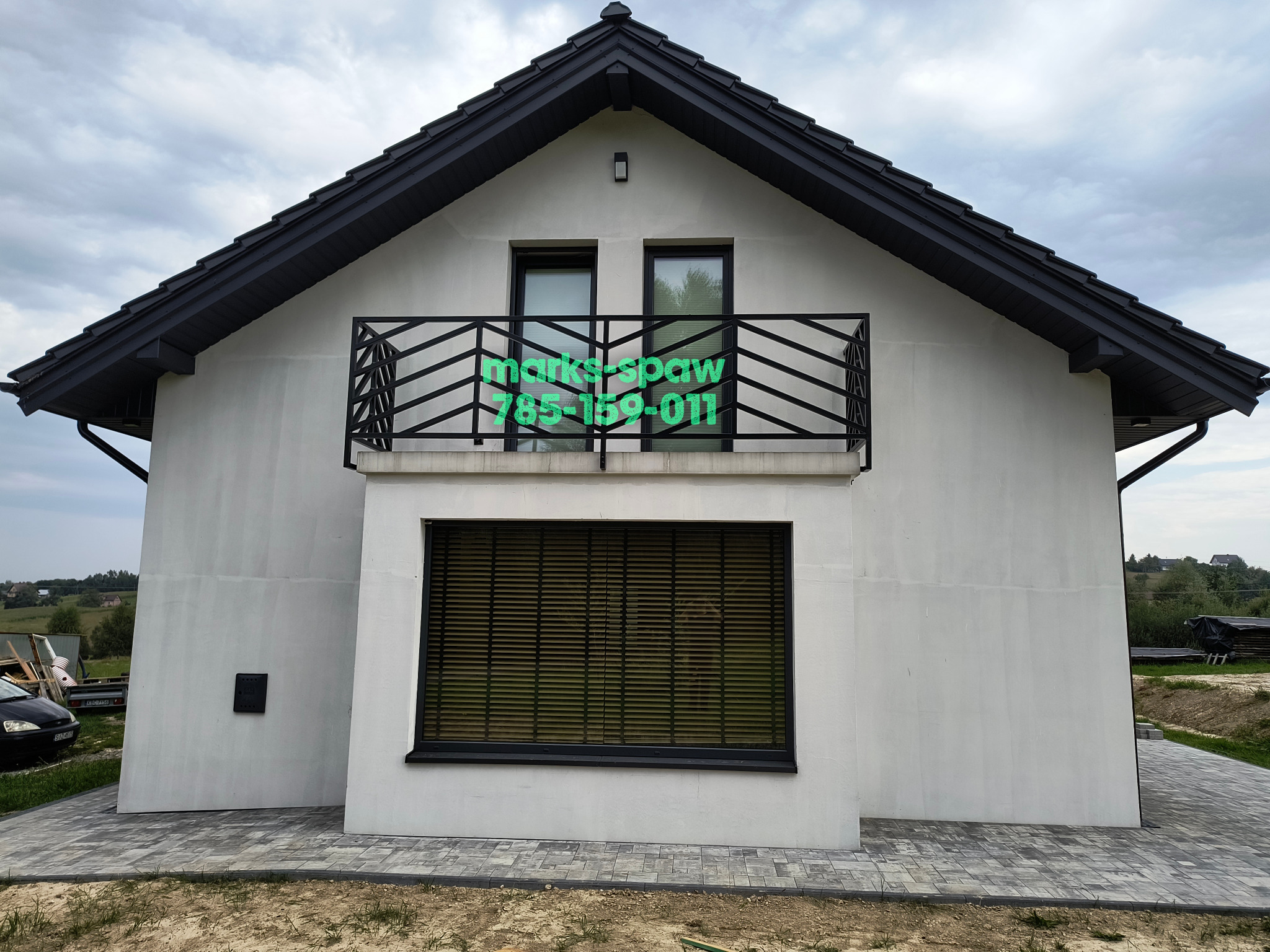 Biały dom z czarnym dachem i balkonem z czarną, nowoczesną balustradą. Na balustradzie zielony napis z nazwą firmy i numerem telefonu.