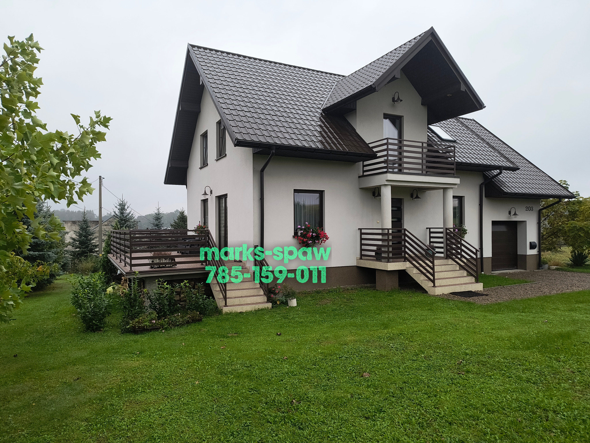 Dom z brązowymi balustradami balkonowymi i tarasowymi, widocznymi zieleń trawnika i kwiaty w oknach. Balustrady metalowe o prostej konstrukcji, dopasowane kolorystycznie do dachu.