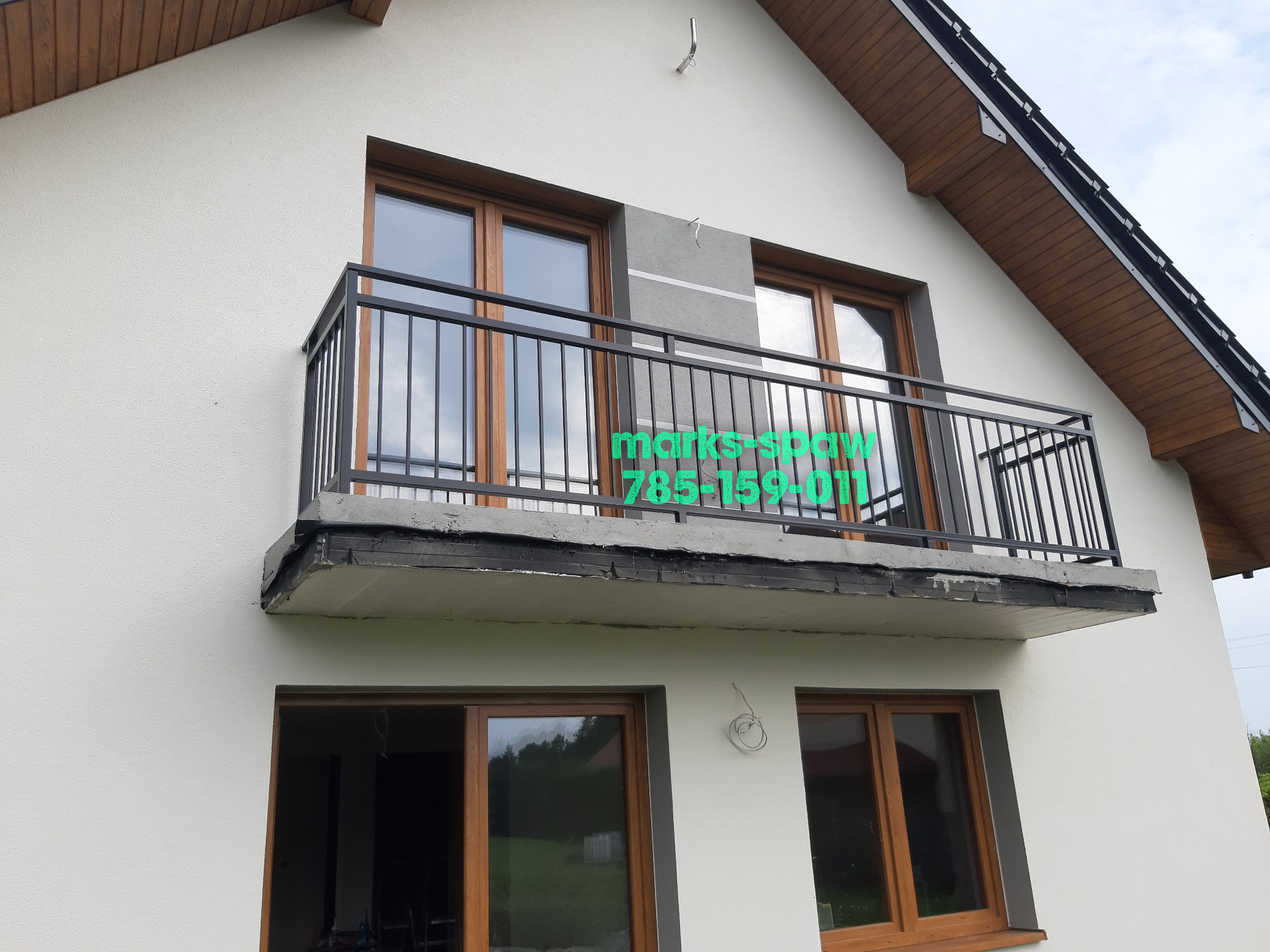 Czarna, metalowa balustrada balkonowa z pionowymi prętami, zamontowana na balkonie domu z drewnianymi oknami. Widoczne detale konstrukcyjne balkonu.