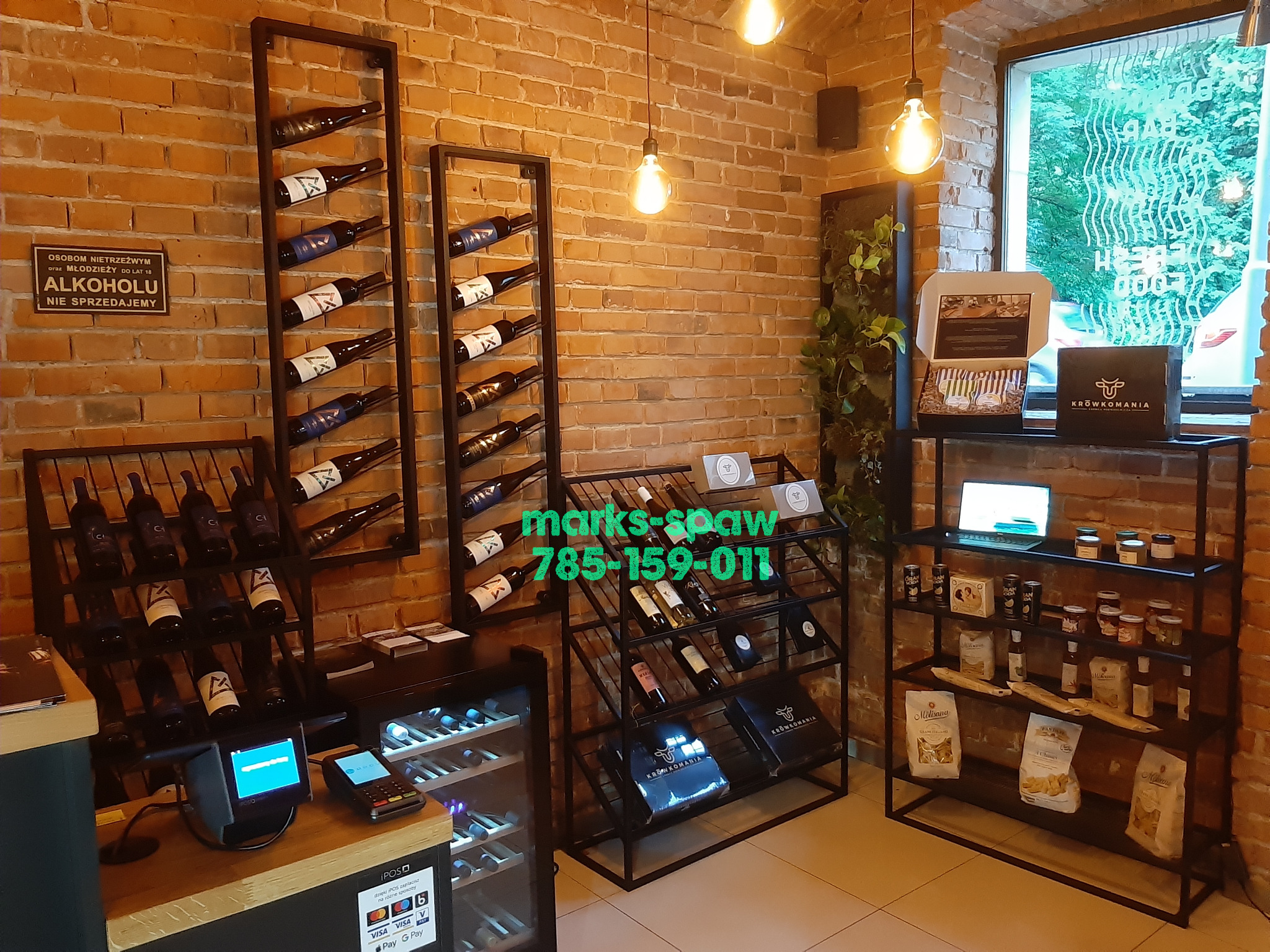 Metalowe regały na wino i produkty spożywcze, z surowego metalu, na tle ceglanej ściany w sklepie, oświetlone żarówkami. Widoczne butelki wina i produkty spożywcze.