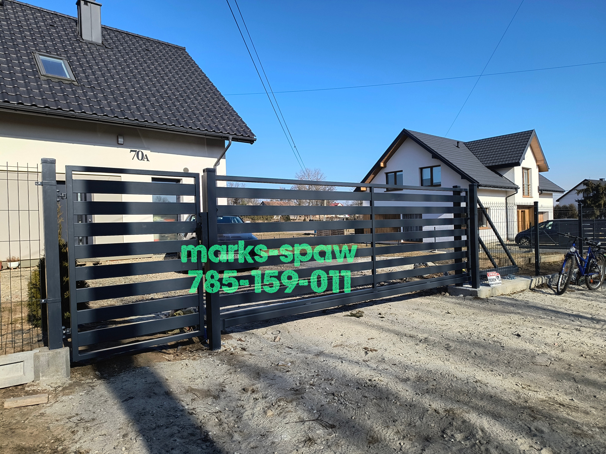 Nowoczesna, pozioma, metalowa brama wjazdowa w kolorze grafitowym, z widocznym numerem telefonu na tle domów jednorodzinnych i błękitnego nieba.