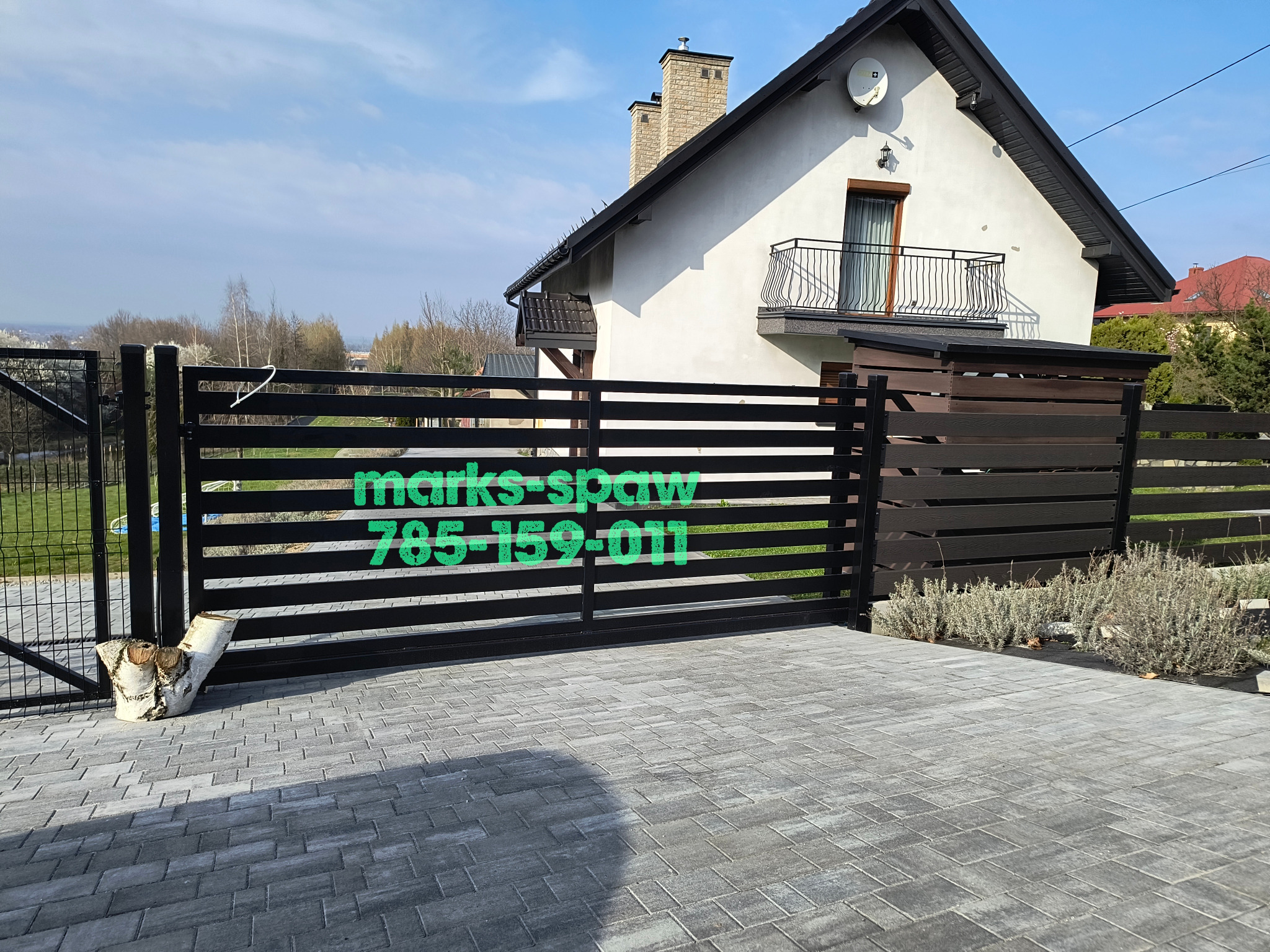 Nowoczesna, pozioma, metalowa brama w kolorze czarnym na tle domu z balkonem. Widoczny fragment ogrodzenia z siatki i kostka brukowa. Na bramie zielony napis.