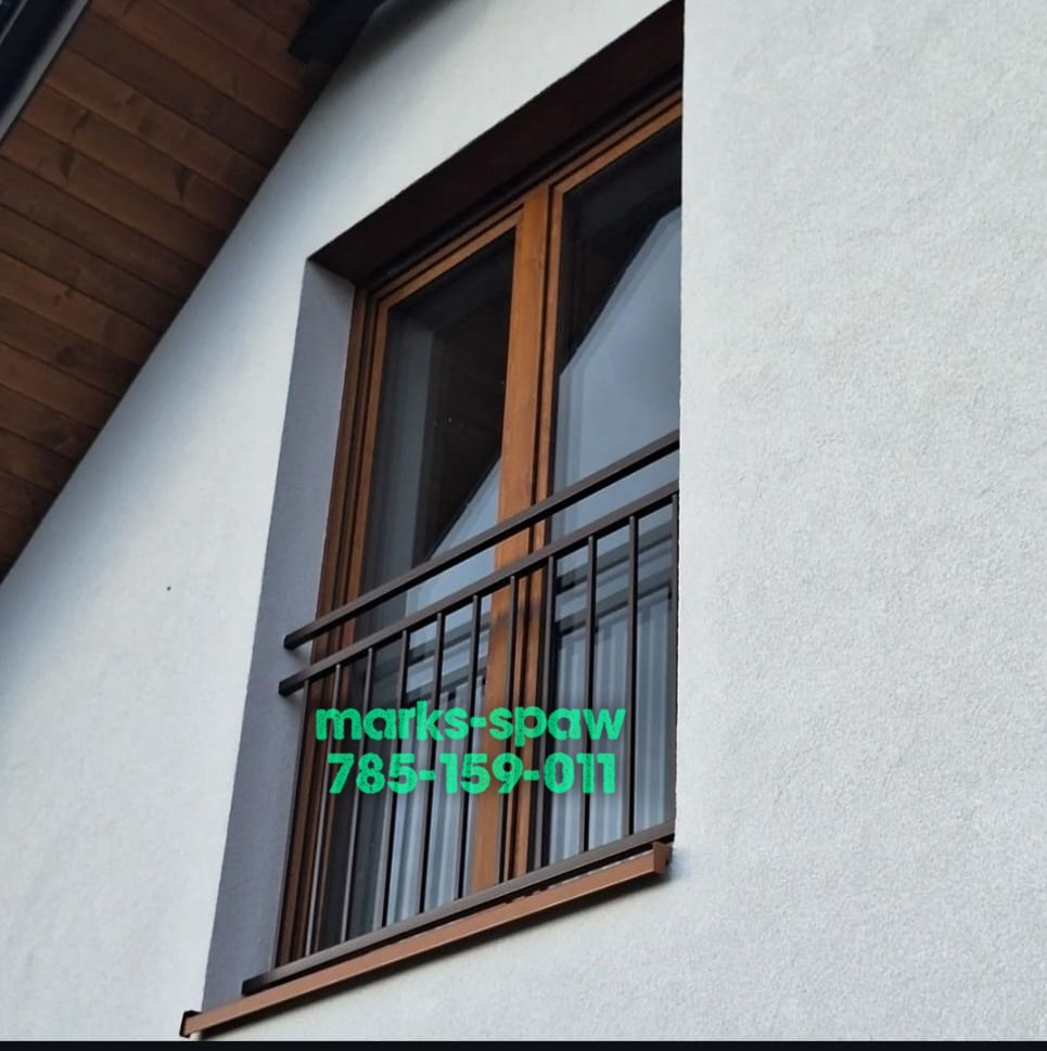 Balkon z brązową, metalową balustradą z pionowymi prętami, zamontowaną przy oknie w budynku z drewnianym podbitką. Widok z dołu, z lekkim kątem.