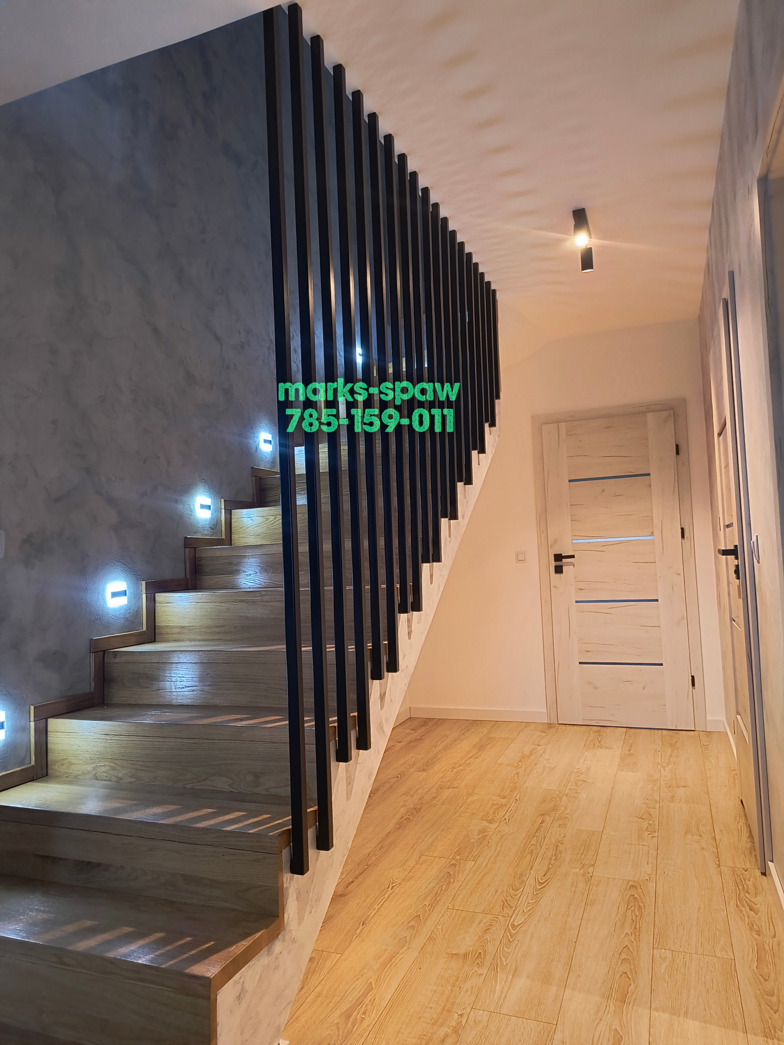 Nowoczesne schody z drewnianymi stopniami, podświetleniem LED i balustradą z pionowych, czarnych prętów. Ściana o fakturze betonu. Widok na korytarz z drewnianą podłogą i drzwiami.