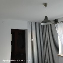 Osa-Bud - Pokój po remoncie: ściana z pionowymi panelami w paski, szara lampa wisząca i biała zasłona przy oknie. Widoczne gniazdka elektryczne i fragment futryny drzwi.