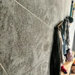 Szara ściana wykończona płytkami imitującymi kamień z jasnymi fugami. Wzdłuż ściany wieszaki z ubraniami i półka. Minimalistyczne wnętrze.