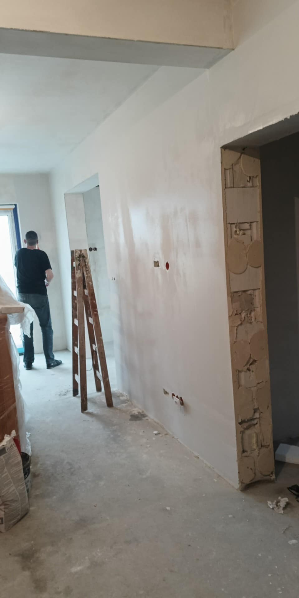 Wnętrze w trakcie szpachlowania ścian, widoczna drabina i pracownik w tle. Ściany pokryte świeżą warstwą gładzi, widoczne puszki elektryczne. Prace wykończeniowe w toku.