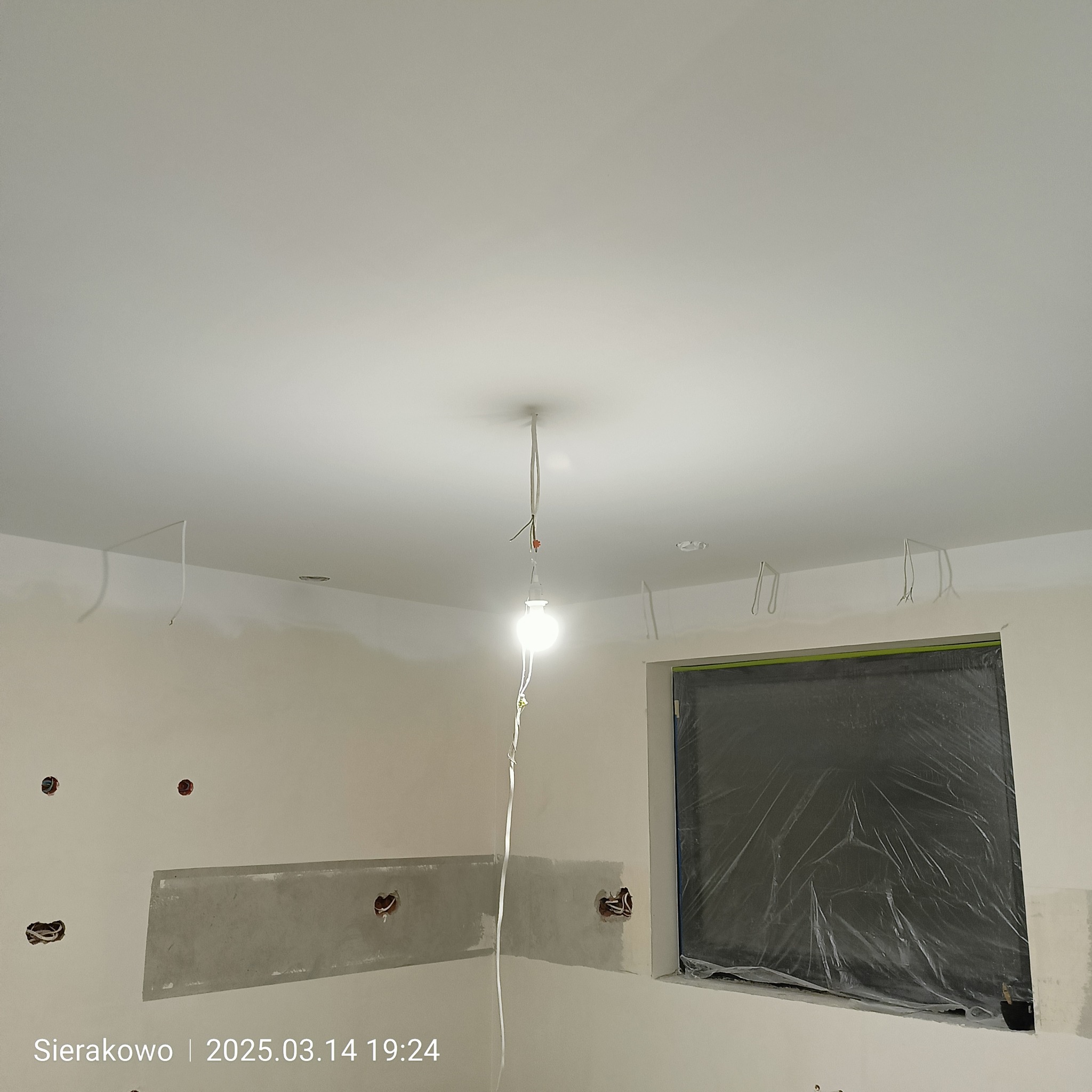 Wnętrze w trakcie remontu: białe ściany, zabezpieczone okno folią, widoczne puszki elektryczne i prowizoryczne oświetlenie. Etap prac wykończeniowych.