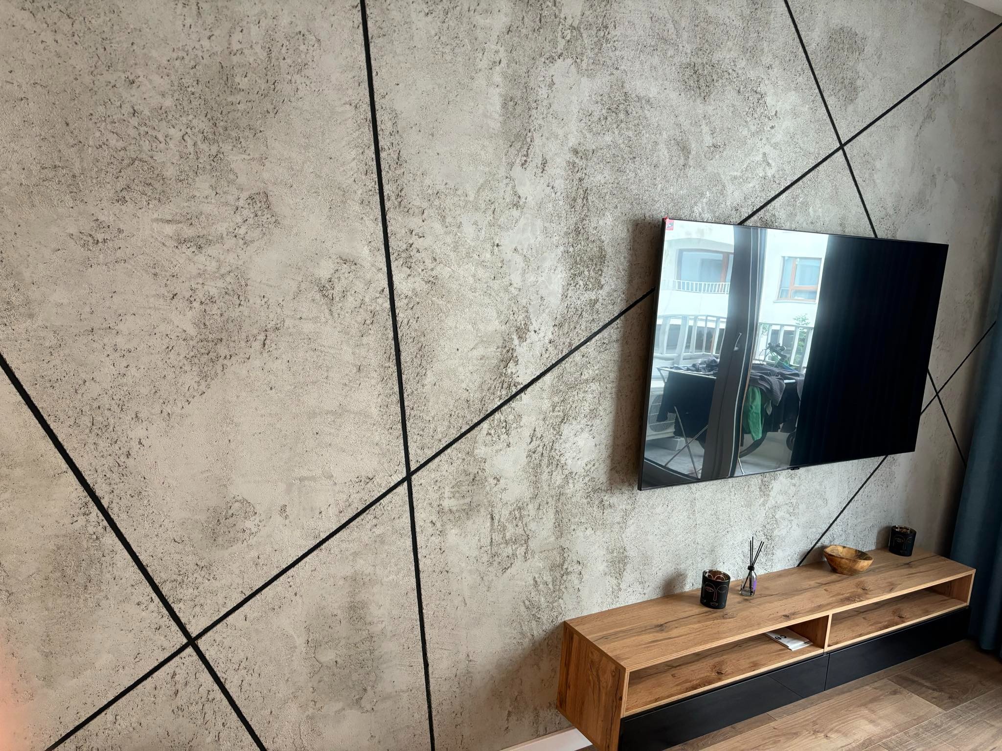 Betonowa ściana z czarnymi, ukośnymi listwami dekoracyjnymi, telewizor i drewniana szafka RTV. Nowoczesny design wnętrza salonu.