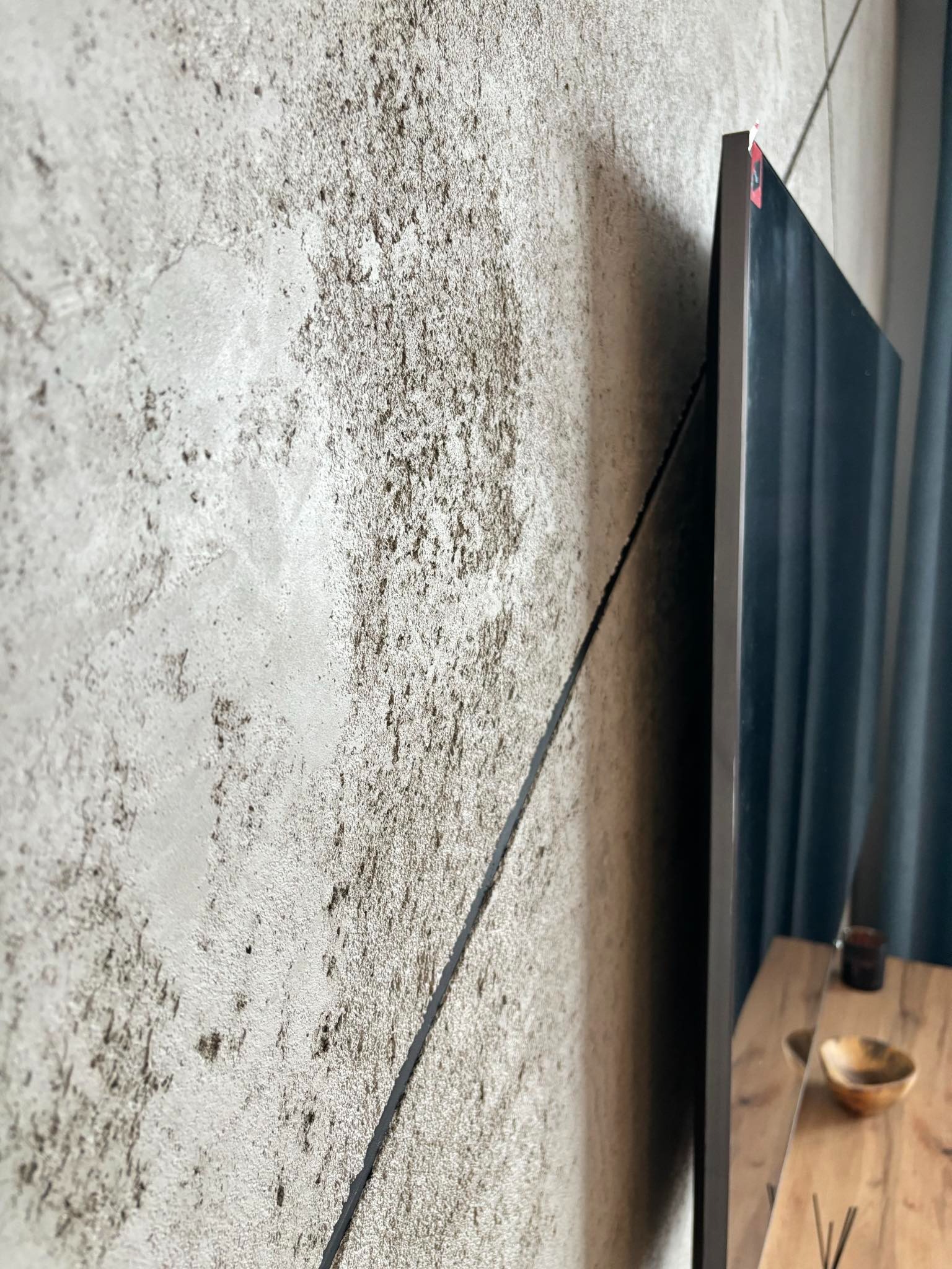 Dekoracyjna ściana z efektem betonu architektonicznego, widoczna z bliska, z fragmentem telewizora i drewnianego stolika. Minimalistyczny design.