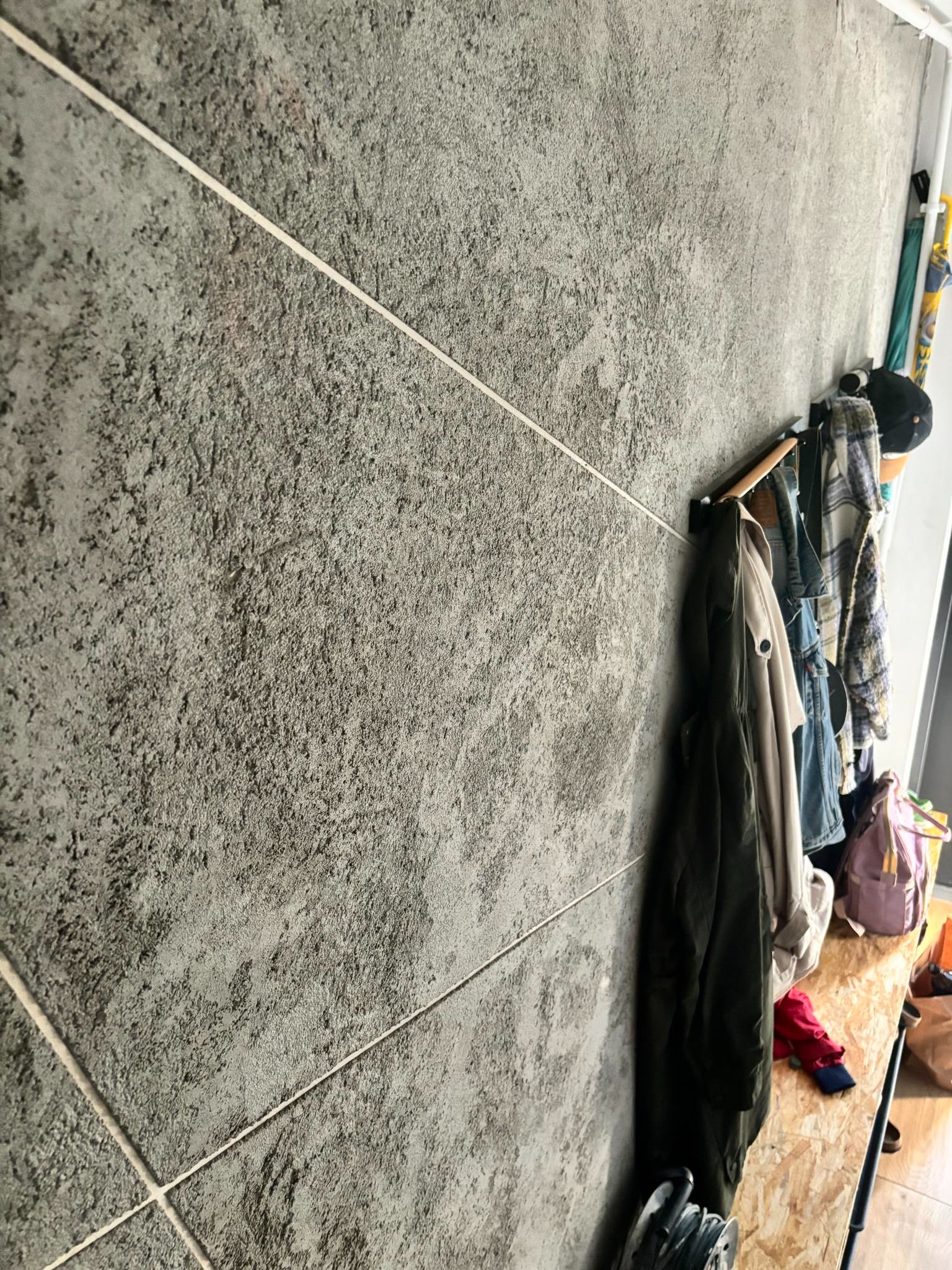 Szara ściana wykończona płytkami imitującymi kamień z jasnymi fugami. Wzdłuż ściany wieszaki z ubraniami i półka. Minimalistyczne wnętrze.