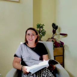 Kobieta w okularach siedzi w fotelu, czytając książkę w jasnym pokoju z roślinami doniczkowymi i obrazem plaży na ścianie. Uśmiech na twarzy, swobodna atmosfera.