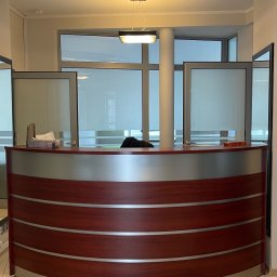 SuperEgo Centrum Psychoterapii Radom - Recepcja z blatem w odcieniach brązu i srebra, widok z przodu. W tle okna z roletami i kolumna. Dwie lampy sufitowe. Podłoga wyłożona płytkami.