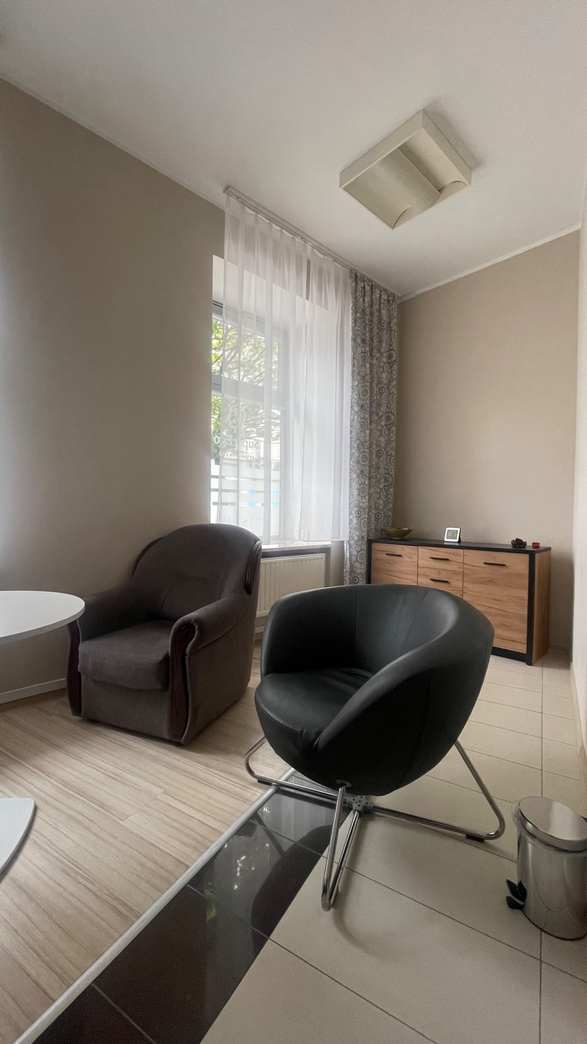 Jasne wnętrze gabinetu z dwoma fotelami, komodą i oknem z firanką. Na podłodze płytki w różnych odcieniach. Neutralna kolorystyka ścian.