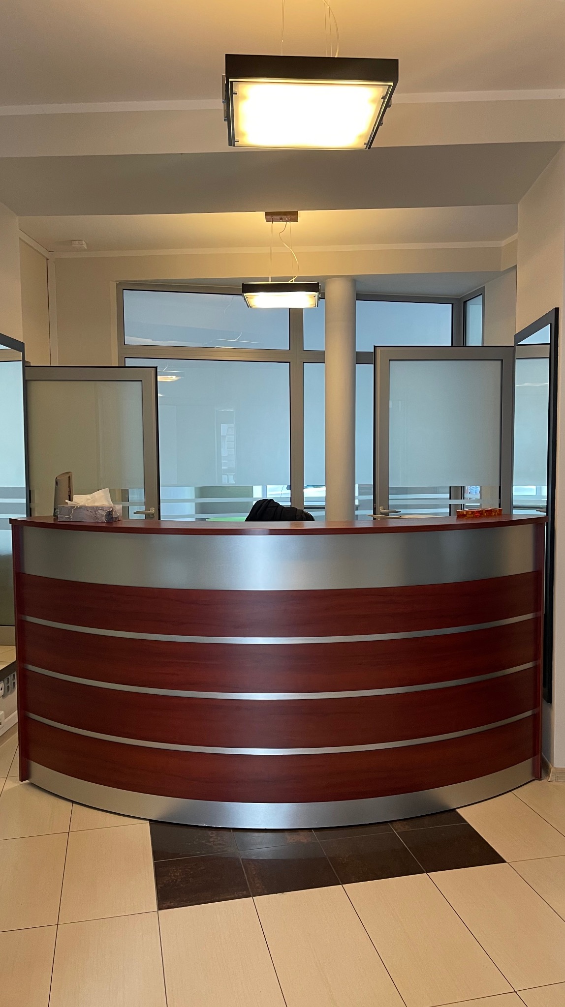 Recepcja z blatem w odcieniach brązu i srebra, widok z przodu. W tle okna z roletami i kolumna. Dwie lampy sufitowe. Podłoga wyłożona płytkami.