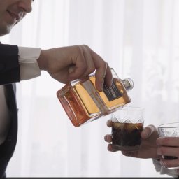 Studio Filmowe Trwaj Chwilo - Elegancki mężczyzna w garniturze nalewa alkohol z markowej butelki do szklanki z lodem, drugi mężczyzna trzyma drugą szklankę. Jasne, stonowane tło.
