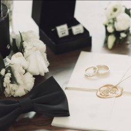 Studio Filmowe Trwaj Chwilo - Eleganckie detale ślubne: perfumy, muszka, spinki, butonierki, obrączki na zapieczętowanej kopercie, tworzące subtelne, stylowe tło dla fotografii ślubnej.