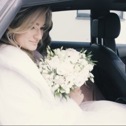 Studio Filmowe Trwaj Chwilo - Panna młoda w sukni ślubnej i welonie siedzi w samochodzie, trzymając bukiet białych kwiatów. Widok z boku na wnętrze pojazdu.