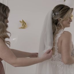 Studio Filmowe Trwaj Chwilo - Panna młoda w koronkowej sukni ślubnej z welonem, poprawianym przez druhnę w jasnoróżowej sukni, w tle dekoracje i balony. Ujęcie z profilu, delikatne światło.