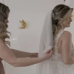 Panna młoda w koronkowej sukni ślubnej z welonem, poprawianym przez druhnę w jasnoróżowej sukni, w tle dekoracje i balony. Ujęcie z profilu, delikatne światło.