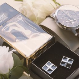 Studio Filmowe Trwaj Chwilo - Elegancki zestaw męski: perfumy Dior, zegarek z brązowym paskiem i spinki do mankietów w pudełku, dopełniony białymi kwiatami. Subtelne oświetlenie podkreśla detale.