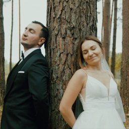 Studio Filmowe Trwaj Chwilo - Para młoda opiera się plecami o drzewo w lesie. Pan młody w czarnym smokingu, panna młoda w białej sukni i welonie. Romantyczna sesja ślubna w naturalnym otoczeniu.