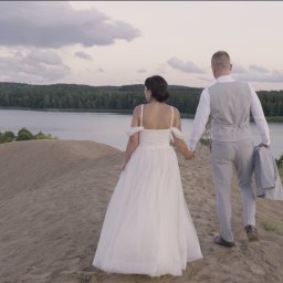 Studio Filmowe Trwaj Chwilo - Para młoda spaceruje po piaszczystym wzgórzu, trzymając się za ręce, z widokiem na jezioro i las w tle, uchwycona w neutralnym oświetleniu.