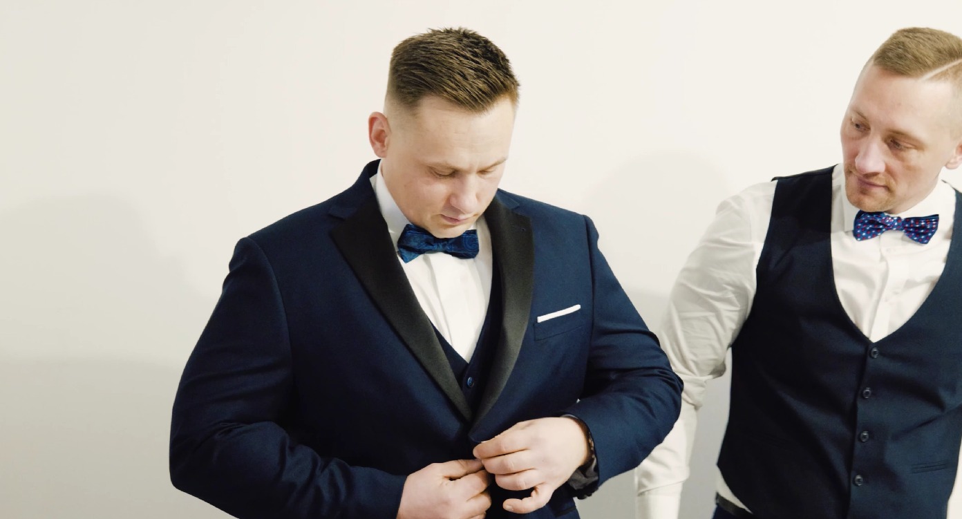 Mężczyzna w granatowym smokingu zapina guzik, obok drugi mężczyzna w kamizelce i muszce. Przygotowania do uroczystości, elegancki strój.