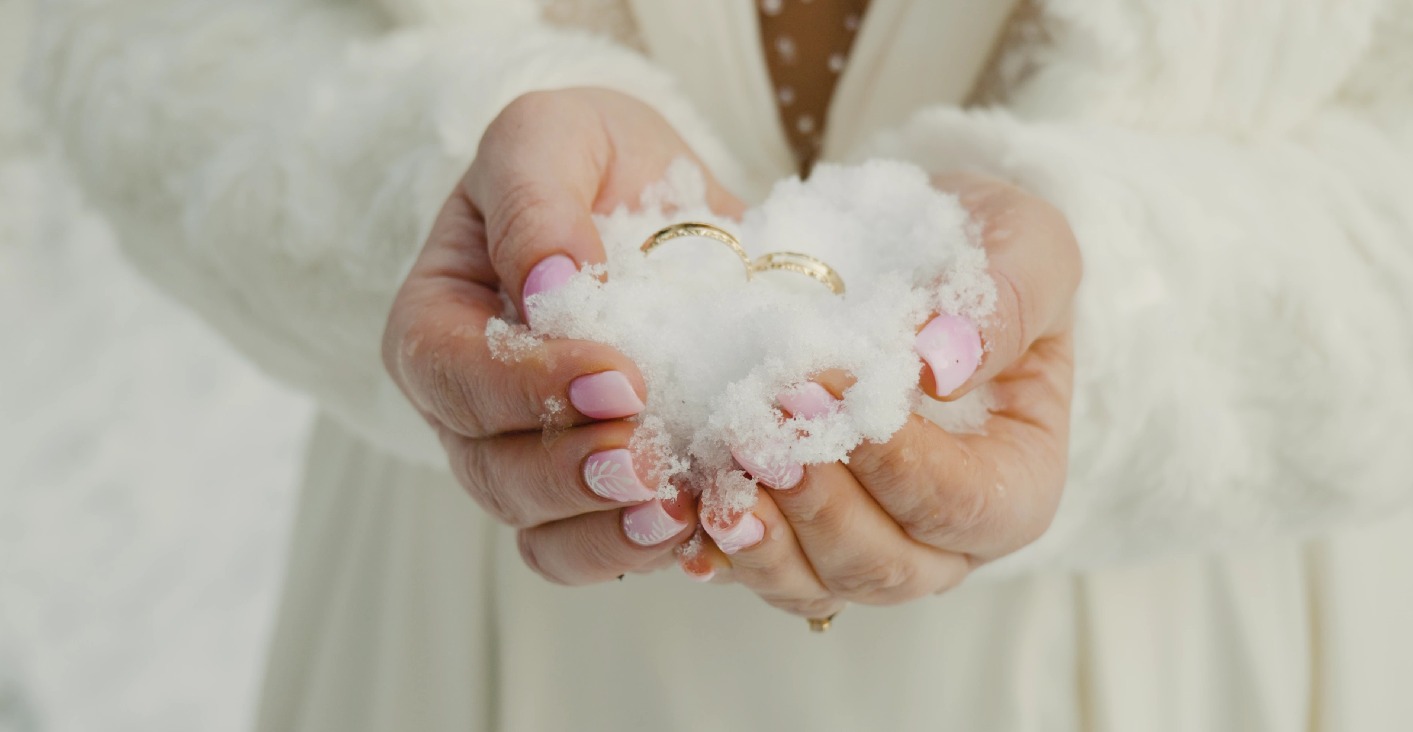 Dłonie panny młodej trzymające śnieg z obrączkami, manicure w odcieniach różu z białymi zdobieniami, białe futerko w tle, delikatne światło.