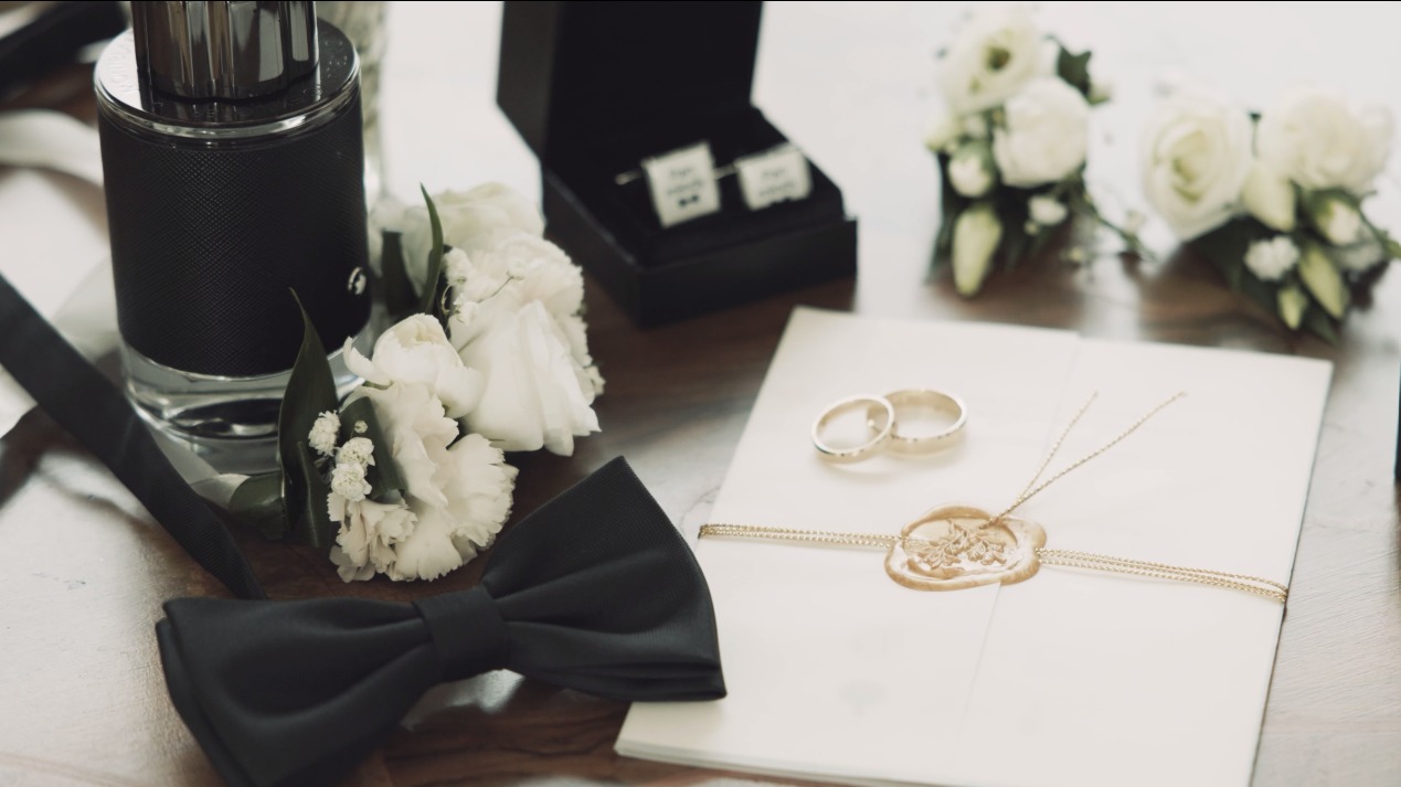 Eleganckie detale ślubne: perfumy, muszka, spinki, butonierki, obrączki na zapieczętowanej kopercie, tworzące subtelne, stylowe tło dla fotografii ślubnej.