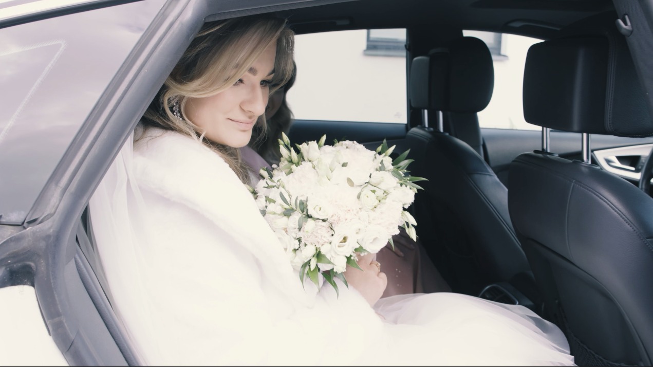 Panna młoda w sukni ślubnej i welonie siedzi w samochodzie, trzymając bukiet białych kwiatów. Widok z boku na wnętrze pojazdu.
