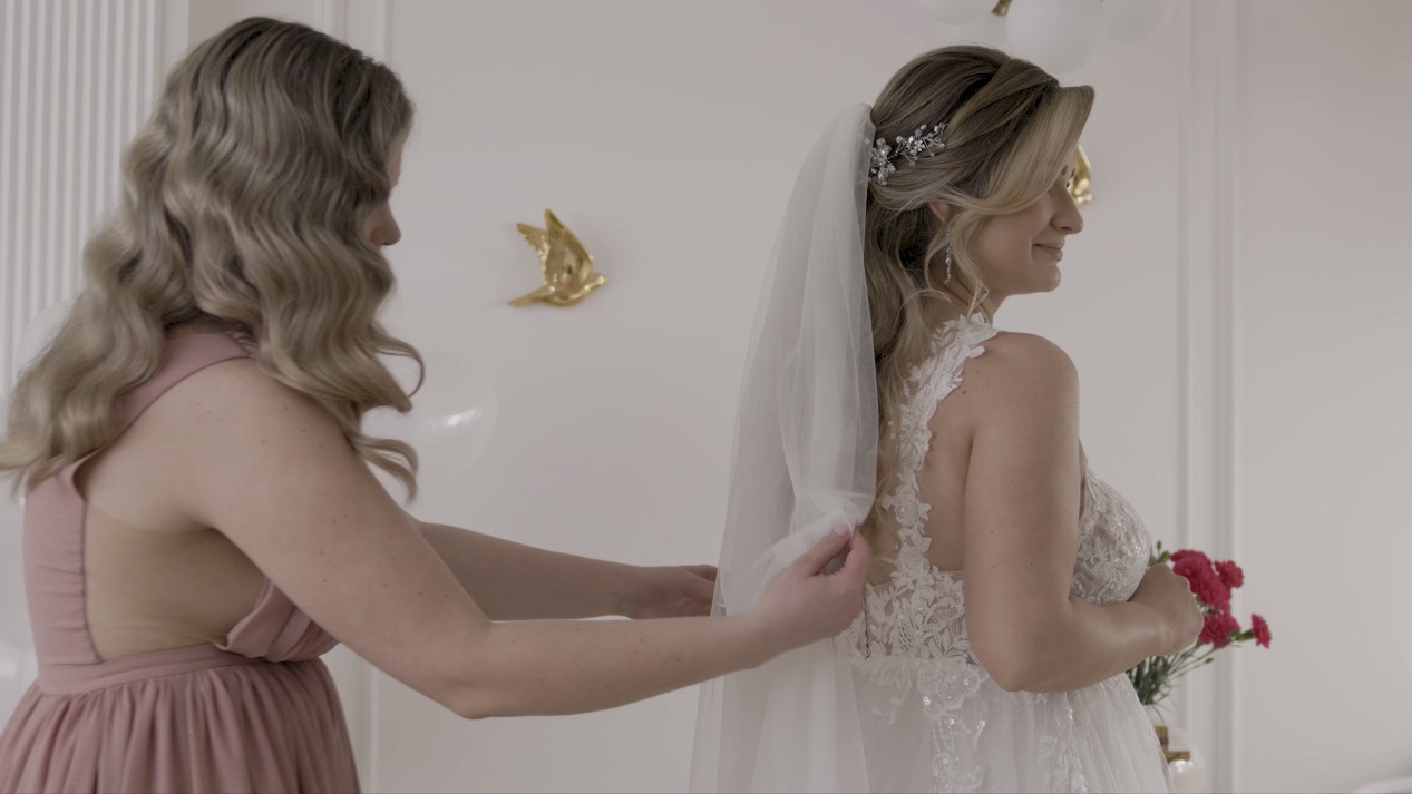 Panna młoda w koronkowej sukni ślubnej z welonem, poprawianym przez druhnę w jasnoróżowej sukni, w tle dekoracje i balony. Ujęcie z profilu, delikatne światło.