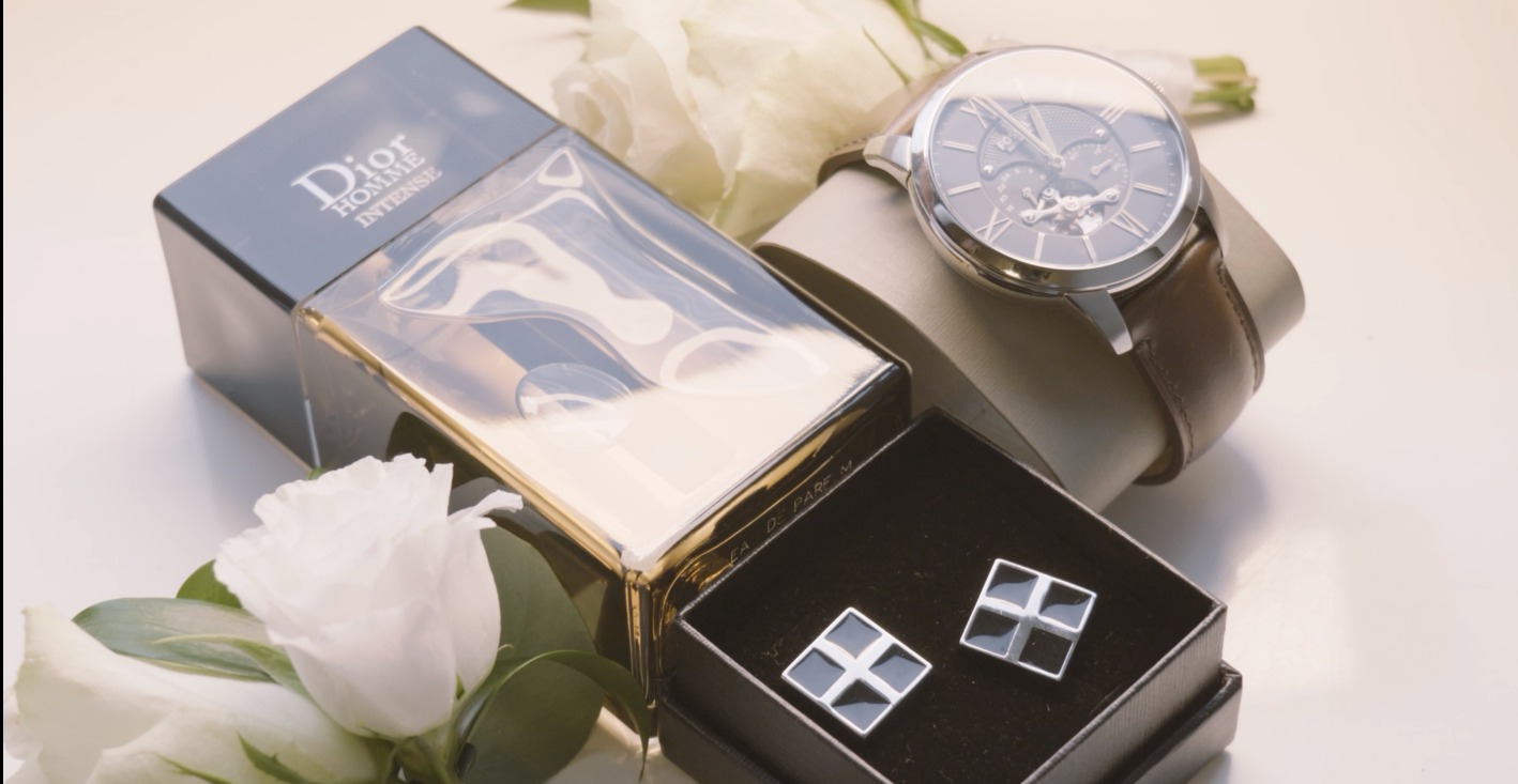 Elegancki zestaw męski: perfumy Dior, zegarek z brązowym paskiem i spinki do mankietów w pudełku, dopełniony białymi kwiatami. Subtelne oświetlenie podkreśla detale.