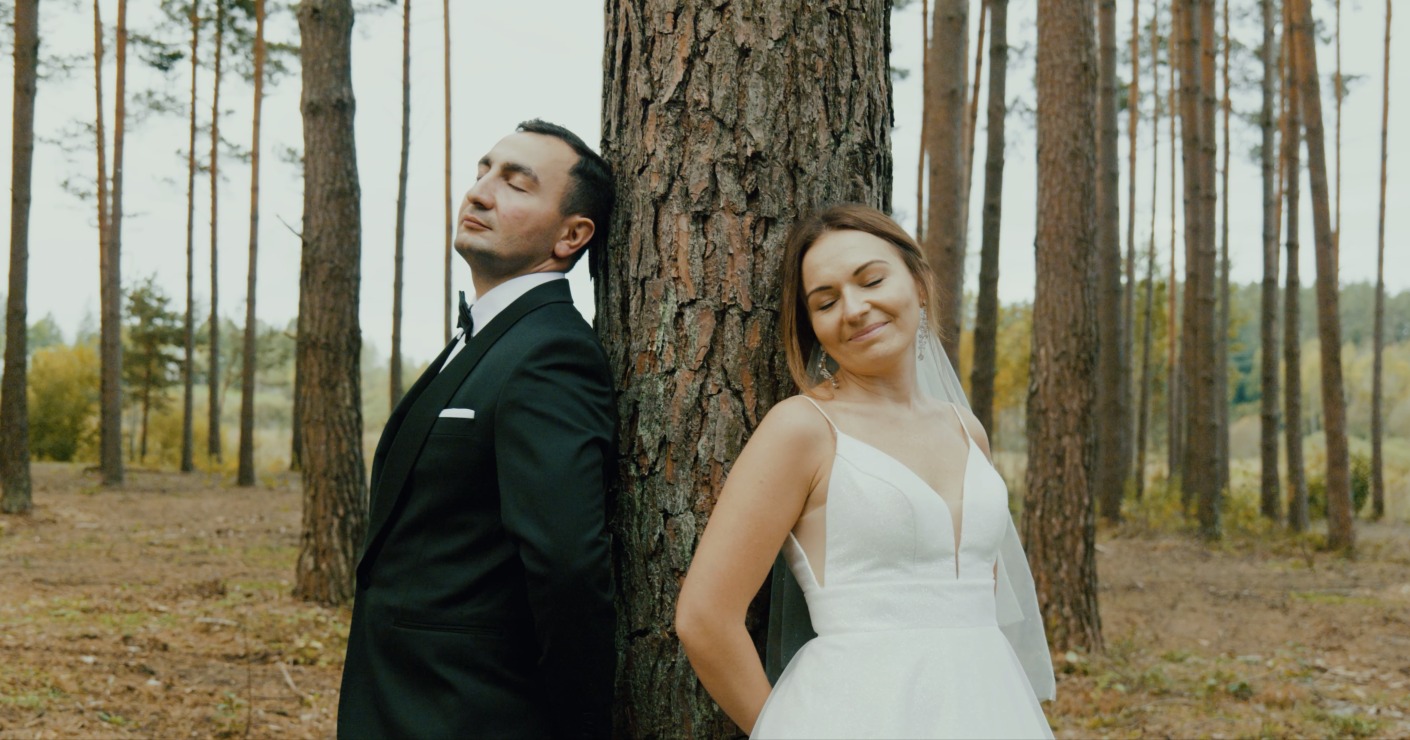 Para młoda opiera się plecami o drzewo w lesie. Pan młody w czarnym smokingu, panna młoda w białej sukni i welonie. Romantyczna sesja ślubna w naturalnym otoczeniu.