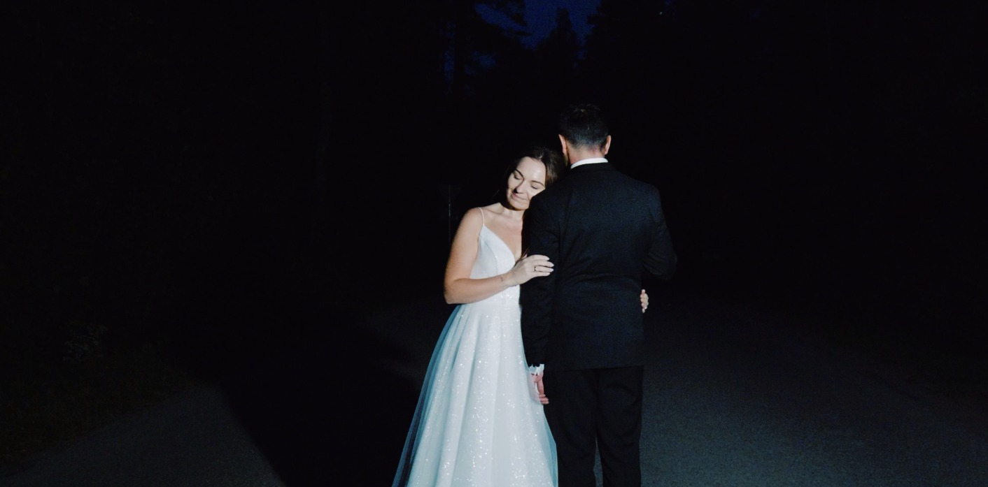 Para młoda w nocnej scenerii: panna młoda w sukni z brokatem opiera się o pana młodego w ciemnym garniturze. Romantyczny kadr w głębokiej czerni.