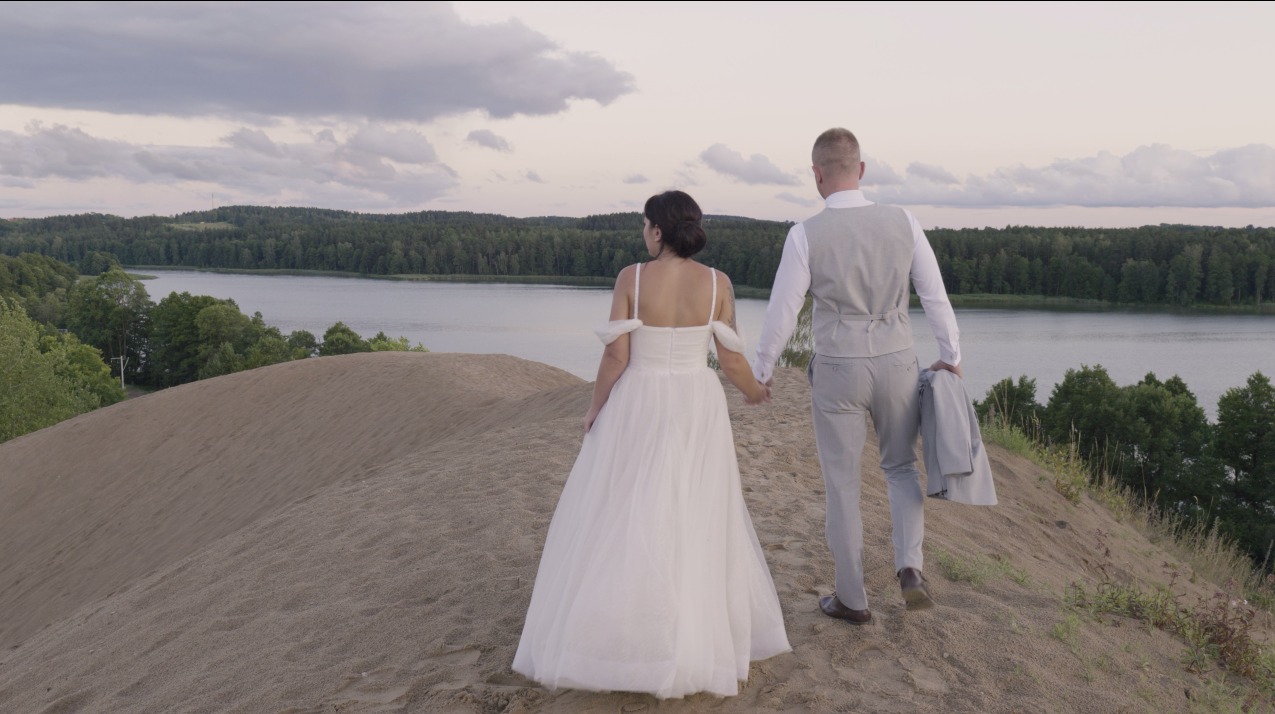 Para młoda spaceruje po piaszczystym wzgórzu, trzymając się za ręce, z widokiem na jezioro i las w tle, uchwycona w neutralnym oświetleniu.
