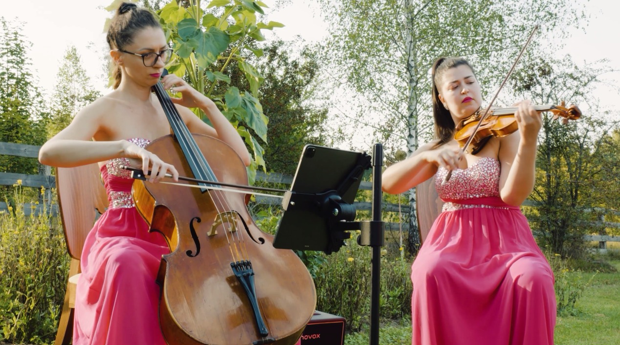 Dwie kobiety w różowych sukniach grające na wiolonczeli i skrzypcach podczas plenerowego występu z nutami na tablecie.