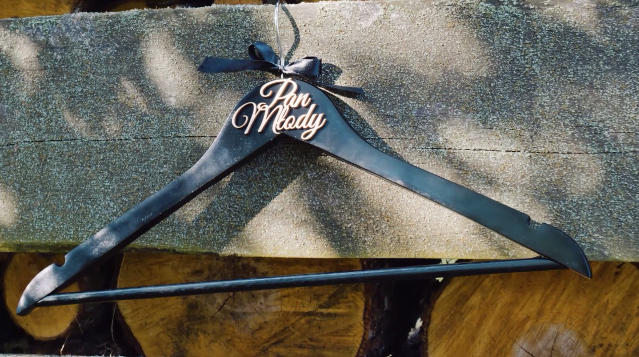 Czarny wieszak z napisem 'Pan Młody' i kokardką, zawieszony na tle drewnianych bali w miękkim świetle. Dekoracyjny detal ślubny, idealny na pamiątkę.